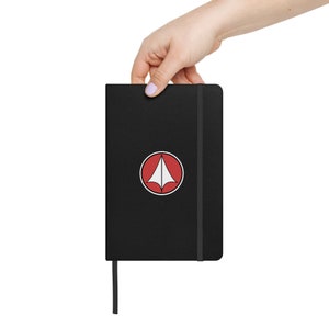 May include: Black notebook with a red circle and white triangle logo. The notebook has a black elastic band closure and a black bookmark.