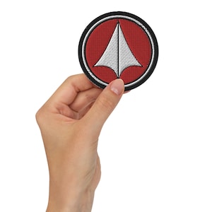 May include: A round, black and red embroidered patch with a white, triangular symbol in the centre. The symbol has a pointed tip and a rounded base.