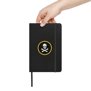 May include: A black notebook with a skull and crossbones design in a yellow circle. The notebook has a black elastic band and a black ribbon bookmark.