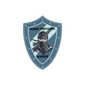 May include: A sticker featuring a shield design with the words "HUMAN TRAFFICKING" and "WARRIOR". The shield has a blue border and a ninja graphic in the center. The ninja is wearing a black head covering and has piercing eyes.