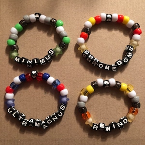 Pode incluir: Quatro pulseiras de contas coloridas com contas de letras pretas que soletram as palavras "MINIMUS", "CHROME", "ULTRAMAGNUS" e "REWIND".