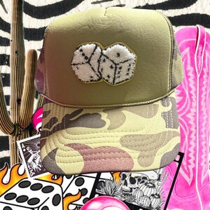 Puede incluir: Gorra de camionero caqui con visera de camuflaje. La gorra presenta un parche de dos dados con textura blanca esponjosa y ribete dorado. Una bota de vaquero rosa y otros gráficos con temática de juego están en el fondo.