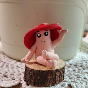May include: A whimsical clay mushroom figure with a red cap and a smiling face, seated on a small wooden slice. The figure has a light pink body and is posed with one arm raised. The background includes a white pot and a crocheted surface.