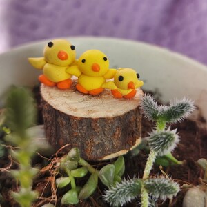 May include: Three handmade yellow clay chicks with orange feet and beaks are perched on a small wooden slice. The chicks have black eyes and outstretched wings. The chicks are in a small planter with green plants and soil.