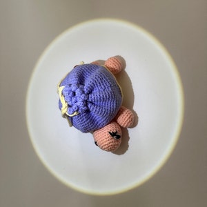May include: A crocheted purple turtle toy with pink legs and a yellow and white crocheted flower on its back.