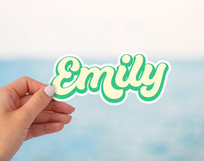 Op de afbeelding: Een groene en gele sticker met de naam "Emily" in een retro-lettertype.