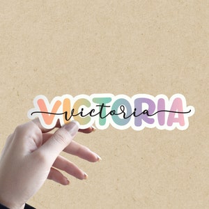 May include: A colorful sticker with the name "VICTORIA" written in a rainbow gradient font. The sticker is on a brown background.