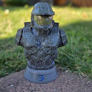 May include: A detailed bust of a Spartan soldier from the Halo video game series. The figure is primarily grey with a pattern of black text covering the helmet and armour. The base is grey with a blue emblem.