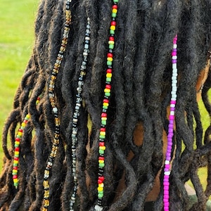 Könnte beinhalten: Vier lange Haarsträhnen, verziert mit bunten Perlen, eingearbeitet in dunkle Dreadlocks. Eine Strähne zeigt ein Muster aus roten, gelben, grünen und schwarzen Perlen. Eine weitere Strähne besteht aus rosa, weißen und violetten Perlen. Zwei zusätzliche Strähnen enthalten transparente, schwarze und bernsteinfarbene Perlen.