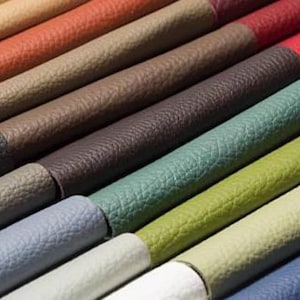 May include: A close-up of a selection of rolled leather samples in various colours. The samples include shades of brown, green, blue, and red. The leather has a textured surface, and the rolls are arranged diagonally.