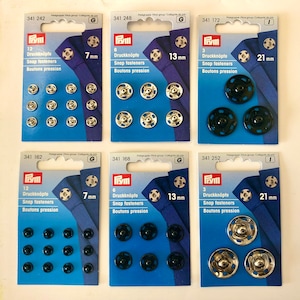 May include: Assortment of six packs of snap fasteners in various sizes. The packs are labelled with the brand name "Frym" and the size of the fasteners in millimetres. The fasteners are silver, black, and gold in colour.