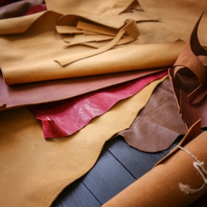 May include: An assortment of leather pieces in various shades of brown, red, and tan, including a rolled piece of leather tied with twine. The leather pieces are arranged on a dark wooden surface.