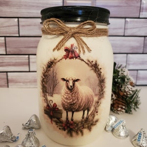 May include: White painted mason jar with a black lid and a burlap bow. The jar has a winter scene with a sheep in a wreath. The jar is surrounded by silver foil wrapped chocolates.