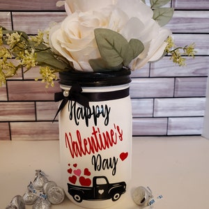 May include: White mason jar vase with a black lid and a black ribbon. The vase is decorated with the words "Happy Valentine's Day" and a red truck with hearts in the bed. The vase is surrounded by silver foil-wrapped chocolates.