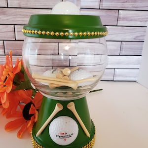 May include: A green and clear glass golf ball holder with gold bead trim. The holder contains golf balls and tees. A golf ball with the text "Pinnacle Power Core 4" is visible.