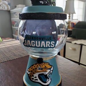 May include: A clear glass gumball machine container featuring a teal base and lid with black glitter accents. The front displays the Jacksonville Jaguars logo and text. A miniature football helmet sits on top of the lid as a decorative topper.