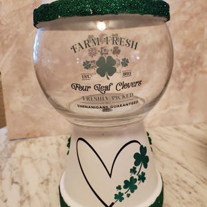 May include: A clear glass jar with a white base and lid, decorated with green glitter and shamrock designs. The jar features the text "Farm Fresh Four Leaf Clovers" and a heart-shaped design. A green glitter ball sits on top.