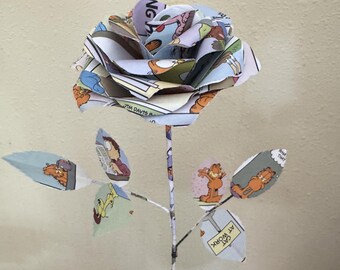 Puede incluir: Una rosa de papel hecha a mano con tiras cómicas de Garfield. La flor presenta una flor de varias capas con hojas y un tallo, mostrando paneles de dibujos animados coloridos. La obra de arte incluye el texto "CAT AT WORK".