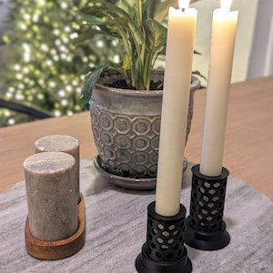 以下が含まれることがあります： ハニカム模様の黒い金属製の燭台に立てられた、白いテーパードキャンドルが2本。キャンドルに火が灯され、燭台は白い大理石の表面に置かれている。背景には緑の葉のついた鉢植えの植物がある。