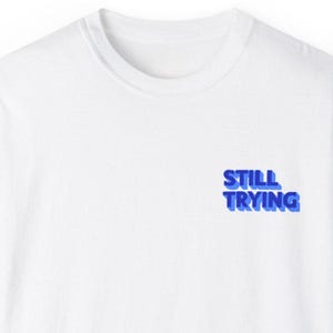 May include: White t-shirt with a blue 3D graphic that says "STILL TRYING" on the front.