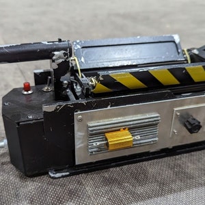May include: A black and silver Ghostbusters proton pack replica with a yellow and black striped design. The pack has a red button, a silver knob, and a gold resistor labeled "2W400".