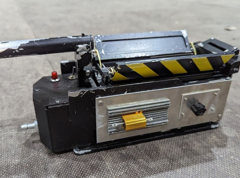 May include: A black and silver Ghostbusters proton pack replica with a yellow and black striped design. The pack has a red button, a silver knob, and a gold resistor labeled "2W400".