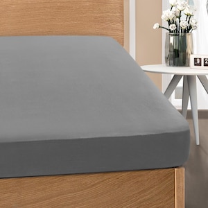 May include: A fitted gray bed sheet on a wooden bed frame. The sheet is a solid color and appears to be made of a soft material. The bed is next to a small white table with a vase of white flowers.