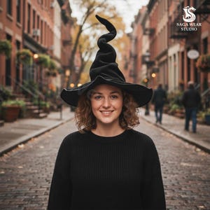 May include: A black witch hat with a tall, curled tip and wide brim. The hat is worn by a person wearing a black sweater. The background shows a brick-paved street with buildings on either side.
