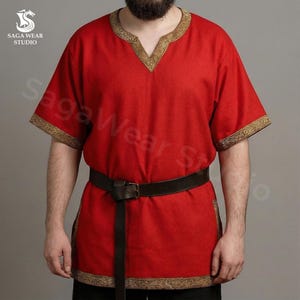 May include: A red tunic with short sleeves and gold trim around the neckline and sleeves. A brown leather belt is worn around the waist. The tunic is part of the Saga Wear Studio collection.