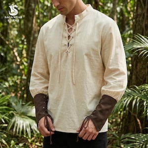 May include: Cream-coloured long-sleeved shirt with a lace-up neckline and brown cuffs. The shirt features a mandarin collar and laced cuffs. The logo "Saga Wear Studio" is visible in the upper left corner.