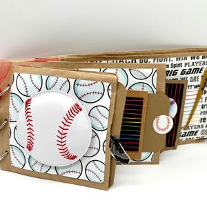 May include: A handmade baseball-themed scrapbook. The cover features a large white baseball with red stitching, set against a background of baseballs. The pages are bound with metal rings and a red ribbon.