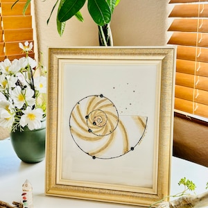 May include: A framed artwork featuring a gold and black spiral design on a white background. The spiral is surrounded by small black dots.