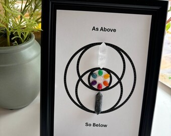 May include: A framed print with a black and white geometric design featuring two overlapping circles and a vertical line. The print includes the text "As Above" and "So Below". The design is accented with small colorful stones and two crystal points.