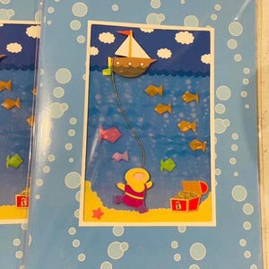 May include: A greeting card featuring a layered paper cutout design of an underwater scene. A brown boat floats on the surface, connected by a string to a diver in a yellow suit swimming near a red treasure chest filled with gold on the sandy floor.