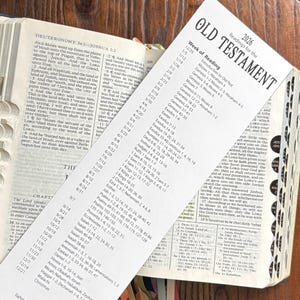 May include: Open Bible with a reading schedule for 2026. The book is open to the book of Deuteronomy, with a reading guide inserted. The pages are cream-colored, and the text is black.