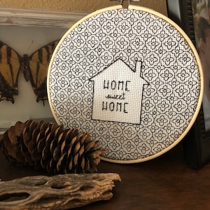 Könnte beinhalten: Ein weißer Stickrahmen mit einem schwarz-weißen geometrischen Muster. In der Mitte des Rahmens befindet sich ein schwarz-weißes Haus mit dem Text "Home sweet Home" im Inneren.