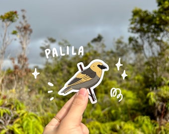 May include: A sticker depicting a Palila bird illustration, perched on a branch. The bird has a yellow head, a black beak, and gray and yellow body. The word "PALILA" is written above the bird in white. The background is a blurred outdoor scene.