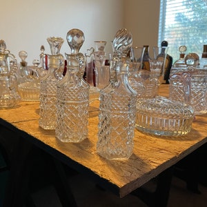 May include: A collection of vintage glass decanters with intricate cut glass designs. The decanters are arranged on a wooden table, with some of them grouped together and others spread out.
