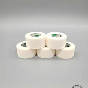 Peut inclure: Six rouleaux de ruban adhésif médical blanc avec une marque 3M verte. Le ruban est empilé en forme de pyramide.