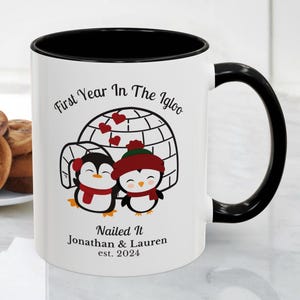 Puede incluir: Taza de cerámica blanca con interior y asa negros. La taza presenta una ilustración de dibujos animados de dos pingüinos frente a un iglú, con el texto "First Year In The Igloo" y "Nailed It Jonathan & Lauren est. 2024".