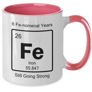 May include: White ceramic mug with a pink rim and handle. The mug features a periodic table element graphic for iron, with the text "6 Fe-nomenal Years", "26", "Fe", "Iron", "55.847", and "Still Going Strong".