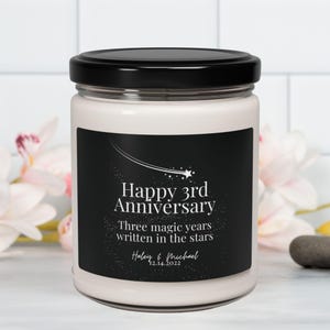 May include: A white candle in a glass jar with a black lid. The label features the text "Happy 3rd Anniversary" with the words "Three magic years written in the stars" and the names "Haley & Michael" with the date 12.14.2022.