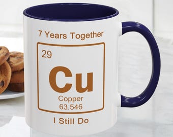 May include: White ceramic mug with a navy blue interior and handle. The mug features the text "7 Years Together" and "I Still Do", along with a copper element design. A plate of cookies is visible in the background.