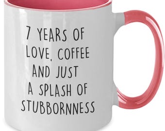 May include: White ceramic mug with a pink interior and handle. The mug features the text "7 YEARS OF LOVE, COFFEE AND JUST A SPLASH OF STUBBORNNESS" in black lettering. A perfect gift for a loved one.