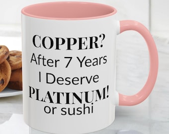 May include: White ceramic mug with a pink interior and handle. The mug features the text "COPPER? After 7 Years I Deserve PLATINUM! or sushi" in black, bold font. A plate of cookies is partially visible in the background.