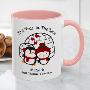 May include: White ceramic mug with a pink interior and handle. The mug features a cartoon illustration of two penguins in front of an igloo, with the text "First Year In The Igloo" and "Nailed It Still Chillin' Together."