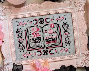 May include: A pink and white cross-stitch embroidery framed in a white ornate frame. The embroidery depicts an open book with a pink and black illustration of a witch's cauldron and a potion bottle. The book is surrounded by a pink and black floral design.