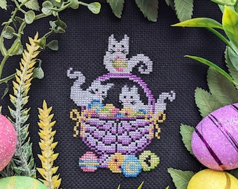 May include: A cross-stitch embroidery design featuring three white cats in a purple basket filled with colorful Easter eggs. The basket is surrounded by green leaves and yellow flowers.