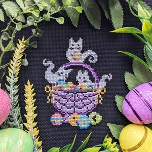 Puede incluir: Un diseño de bordado de punto de cruz con tres gatos blancos en una cesta morada llena de huevos de Pascua coloridos. La cesta está rodeada de hojas verdes y flores amarillas.