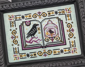 May include: Cross-stitch artwork in a black frame. The design features a raven holding a key, and a caged bird, set against a light blue background. The artwork is framed with decorative borders in black, gold, and purple.